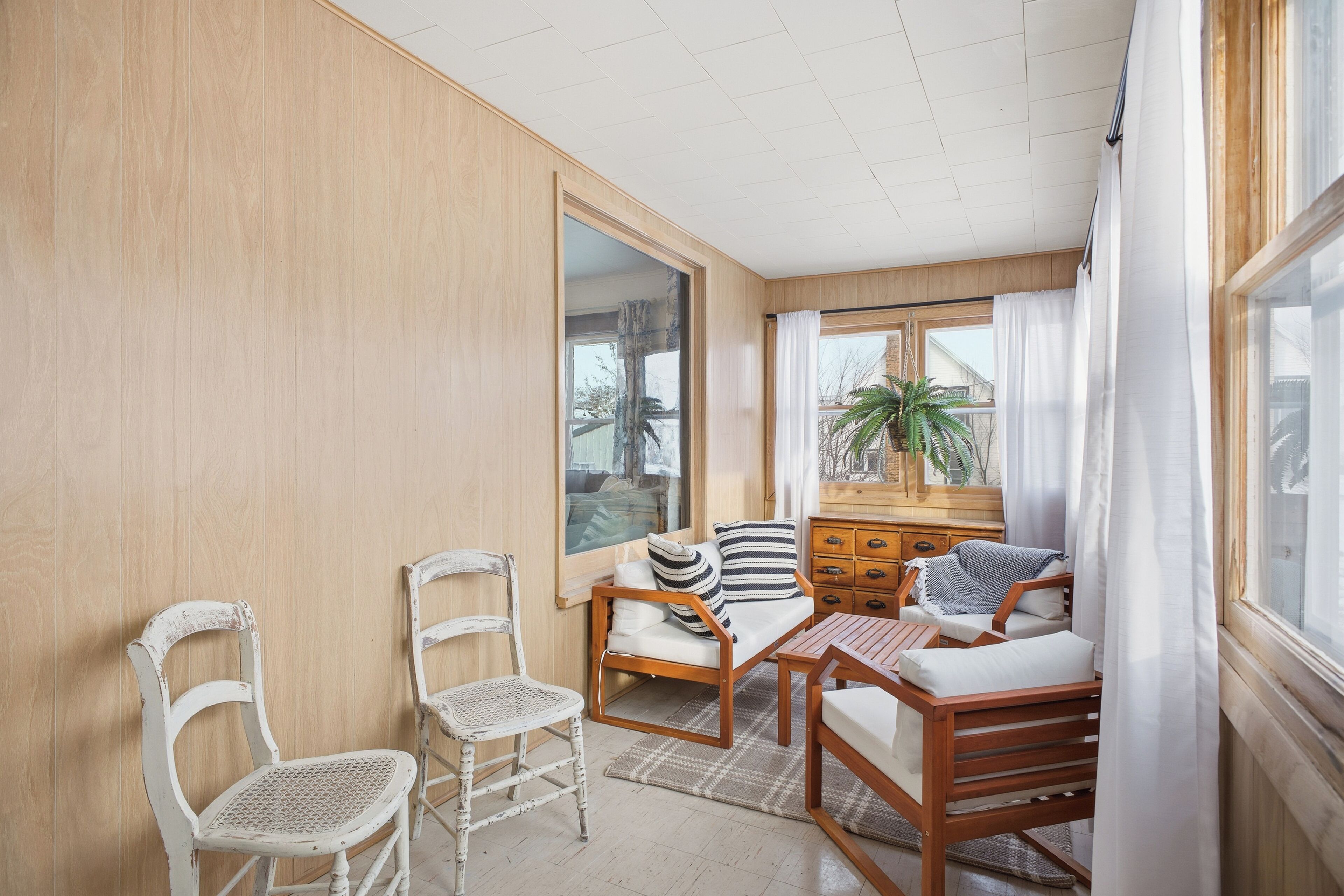 A room with a wooden wall, a sofa, a wooden table, two white chairs, a window with curtains, and a plant.