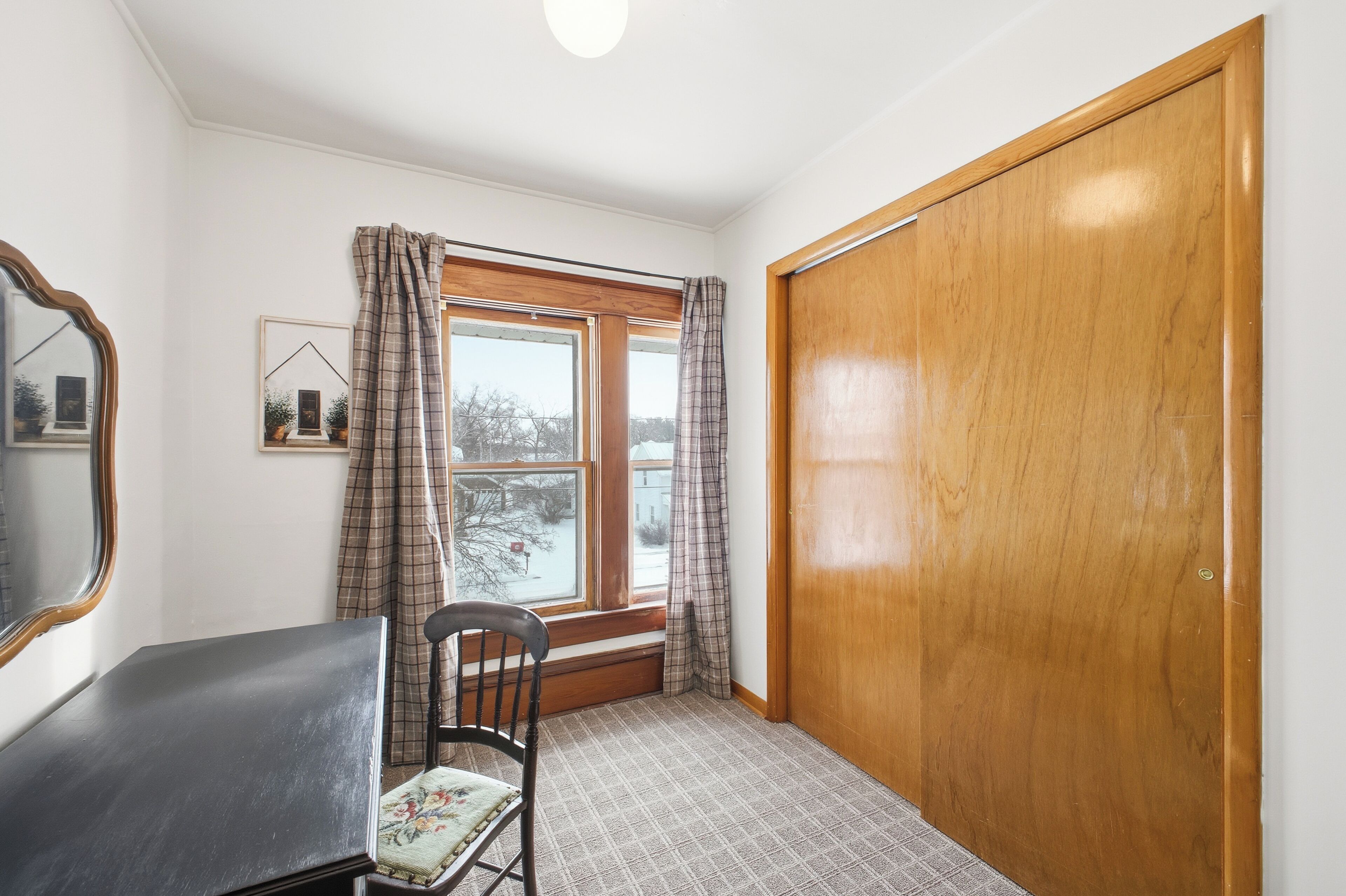 Une pièce avec une porte en bois, une fenêtre ornée de rideaux à carreaux, une chaise et un tableau encadré accroché au mur.