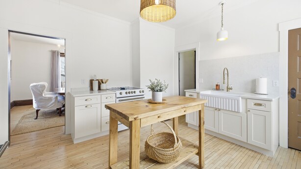 A modern kitchen with white cabinetry, a wooden table, a wicker basket, and a hanging light fixture.