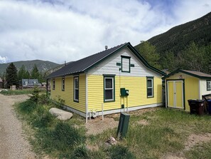 A yellow building with a gabled roof, a small attached shed, and a gravel path leading to it.