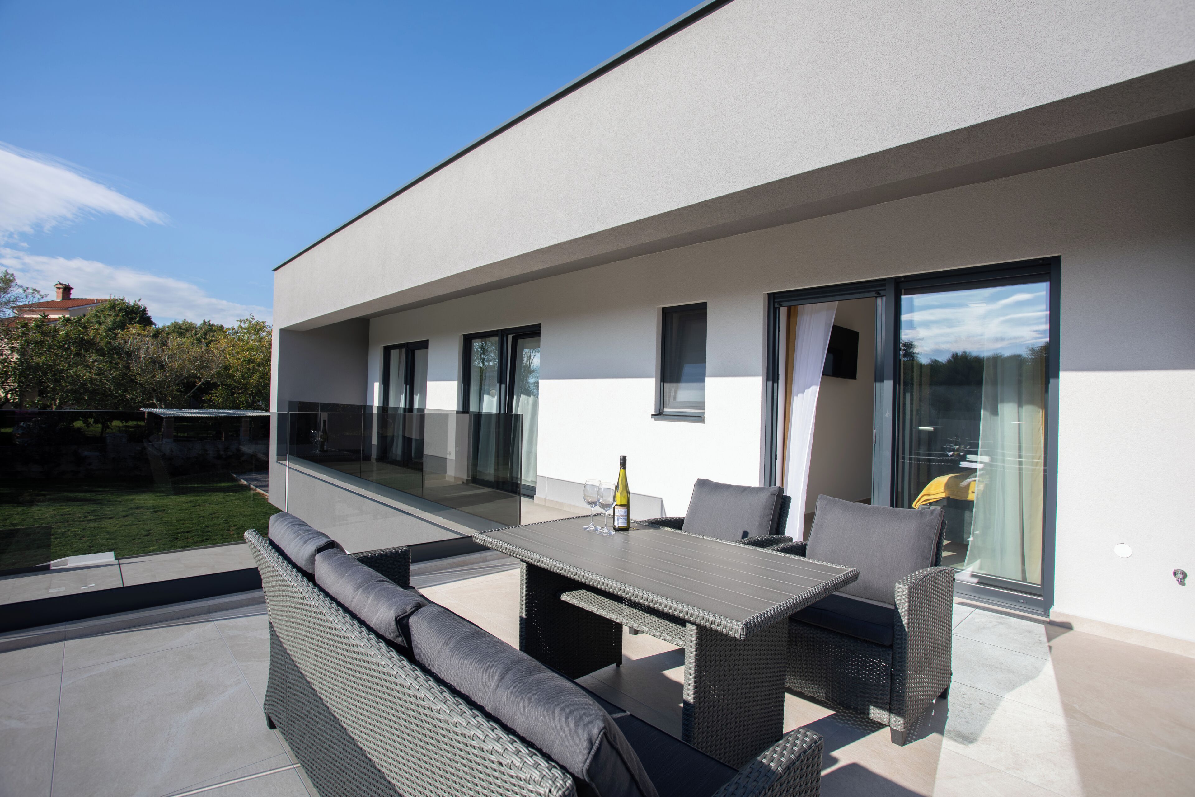 Une terrasse extérieure avec une table et des chaises, une porte vitrée donnant sur un jardin et une façade de bâtiment moderne.