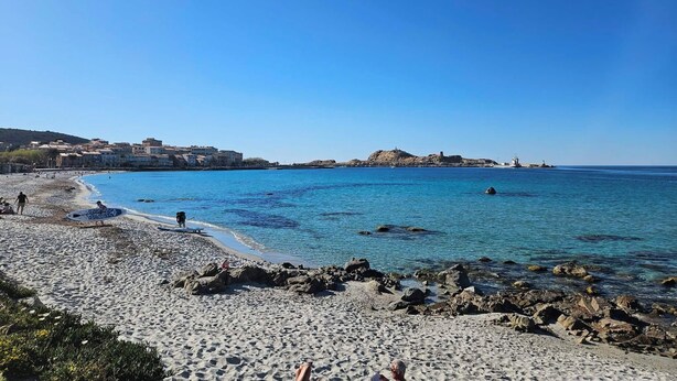 Ein Strand mit Kieselsteinen, blauem Meer und Gebäuden im Hintergrund.