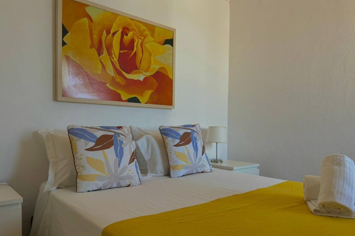 Une chambre d’hôtel avec un lit, des oreillers, une couverture jaune, un tableau, une lampe et une serviette enroulée.