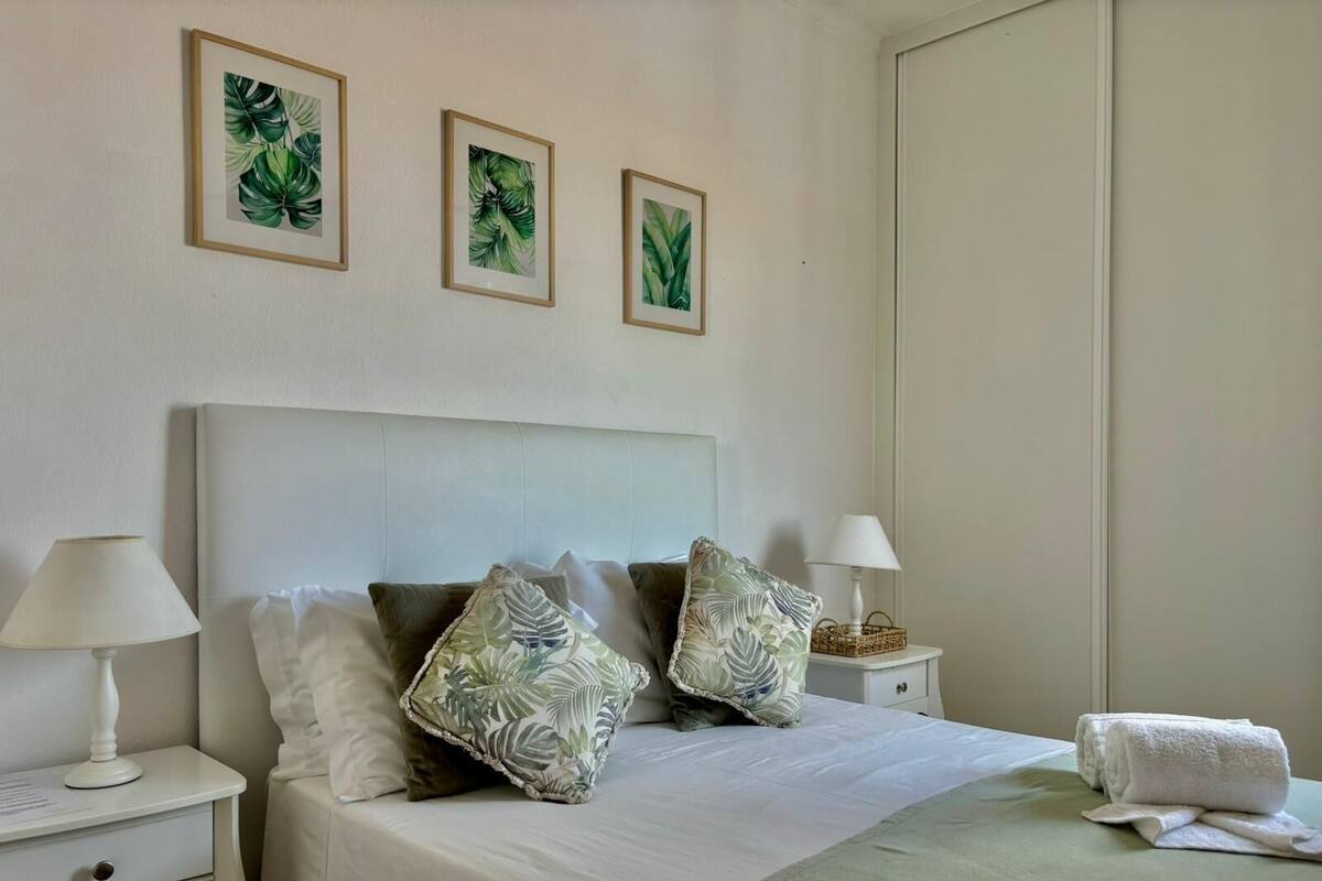 Un lit bien fait, agrémenté d’un plaid vert, deux tables de chevet avec des lampes et trois œuvres d’art représentant des feuilles, encadrées et accrochées au mur.