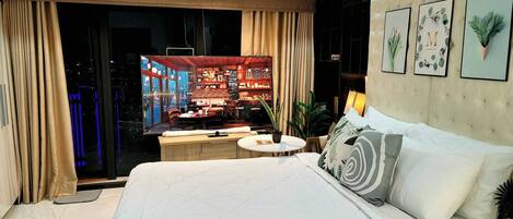 A modern hotel room with a large bed, a small round table, a potted plant, and a view of the hotel's interior.