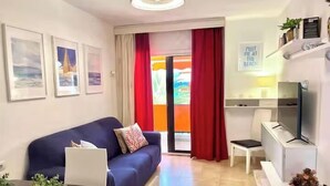Una sala de estar moderna con un sofá, una mesita con una computadora portátil, un televisor y una ventana con cortinas rojas.
