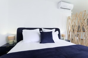 A neatly made bed with white and dark blue pillows, a black headboard, and a white wall with an air conditioning unit. - Studio Sable - Private Jacuzzi - Portbail sur mer center - Swimming pool access (Port-Bail-sur-Mer)