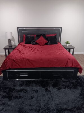 A bed with a black frame, red bedding, and black and red pillows. There is a small table with a lamp and a potted plant beside the bed.
