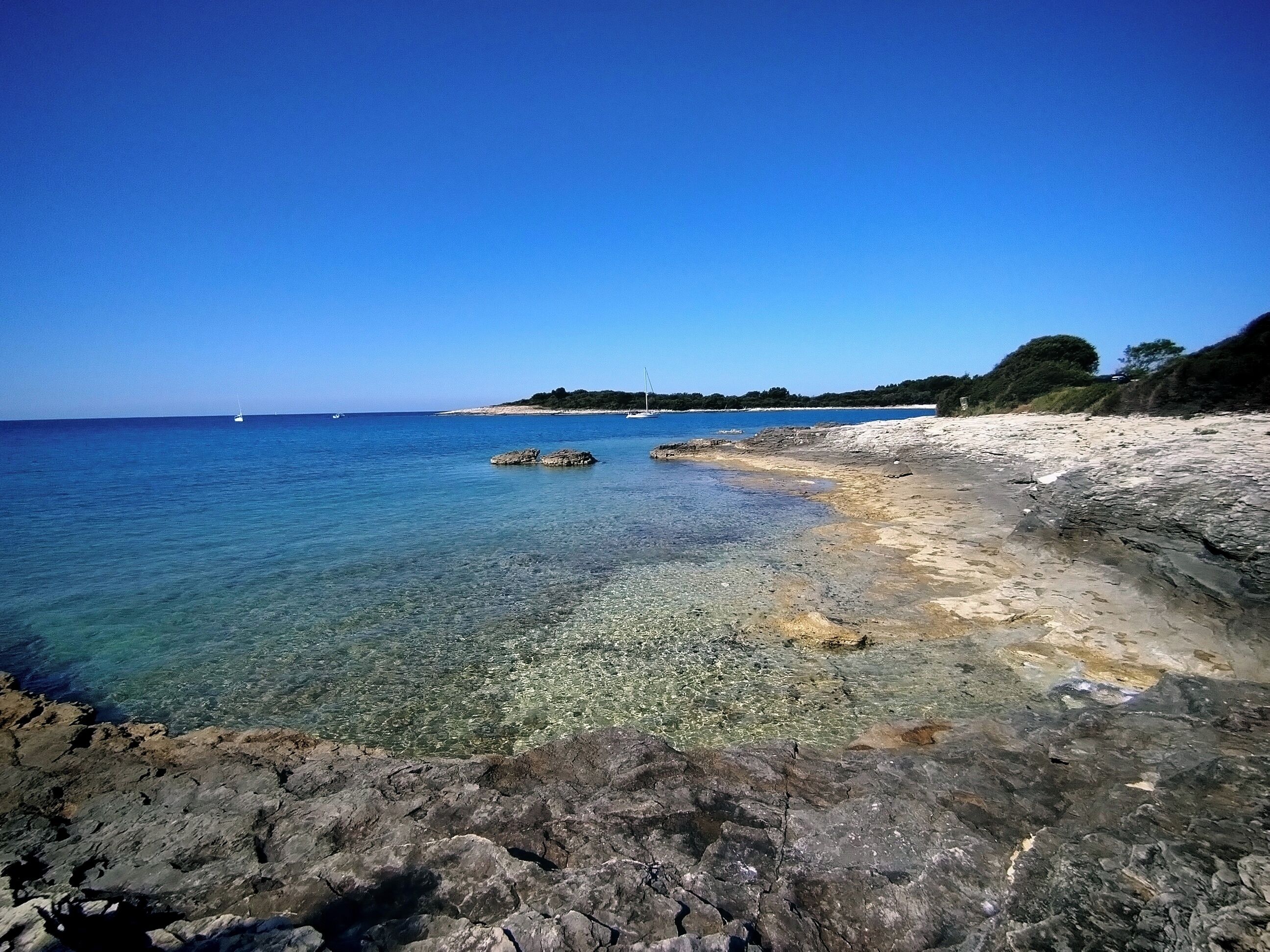 Una playa costera con agua cristalina, costa rocosa y árboles a lo lejos.