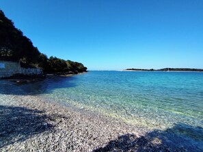 A pebble beach with clear water, a stone wall, and distant land. - Holiday home nearby the sea (Banjole)