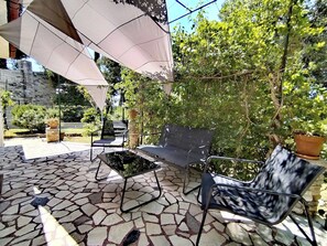 Eine Terrasse mit Steinboden, einem schwarzen Metallstuhl, einer schwarzen Couch und einem weißen Sonnenschirm.