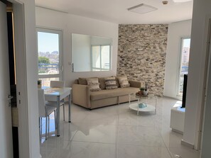A modern living room with a beige sofa, a glass coffee table, and a city view through large windows. - Seafront 1 Bedroom Apartment - Steps from Ashkelon Beach & Marina (Ashkelon)