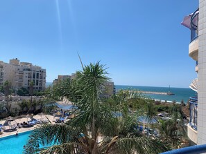 A pool area with lounge chairs and umbrellas, a clear view of the sea, and a building with balconies. - Seafront 1 Bedroom Apartment - Steps from Ashkelon Beach & Marina (Ashkelon)