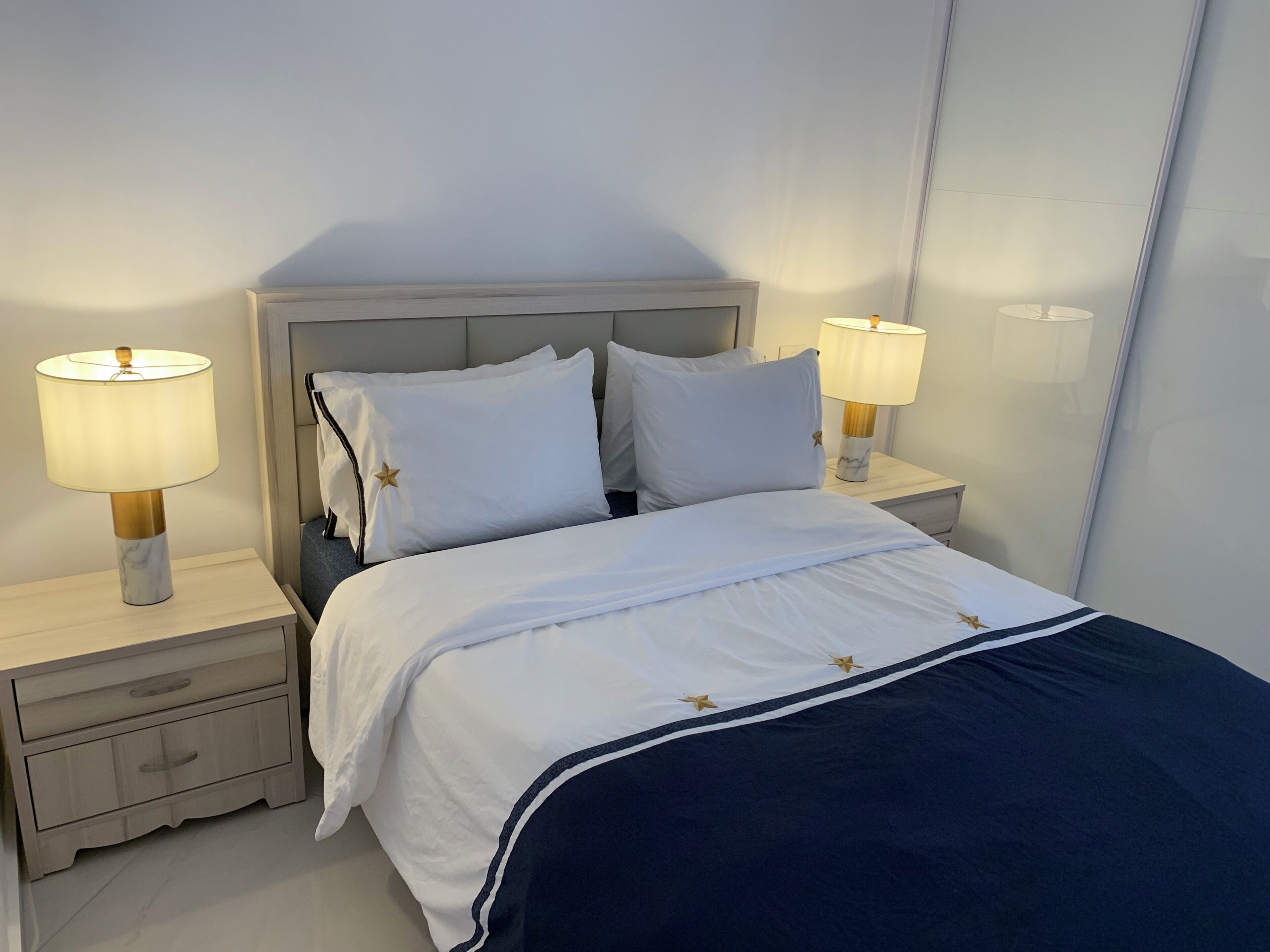 Ein ordentlich bezogenes Bett mit weißer Bettwäsche und einem marineblauen Überwurf, flankiert von zwei Nachttischen mit Lampen, in einem Zimmer mit schlichter Wand und einem Kopfteil.