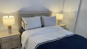 Ein ordentlich bezogenes Bett mit weißer Bettwäsche und einem marineblauen Überwurf, flankiert von zwei Nachttischen mit Lampen, in einem Zimmer mit schlichter Wand und einem Kopfteil.