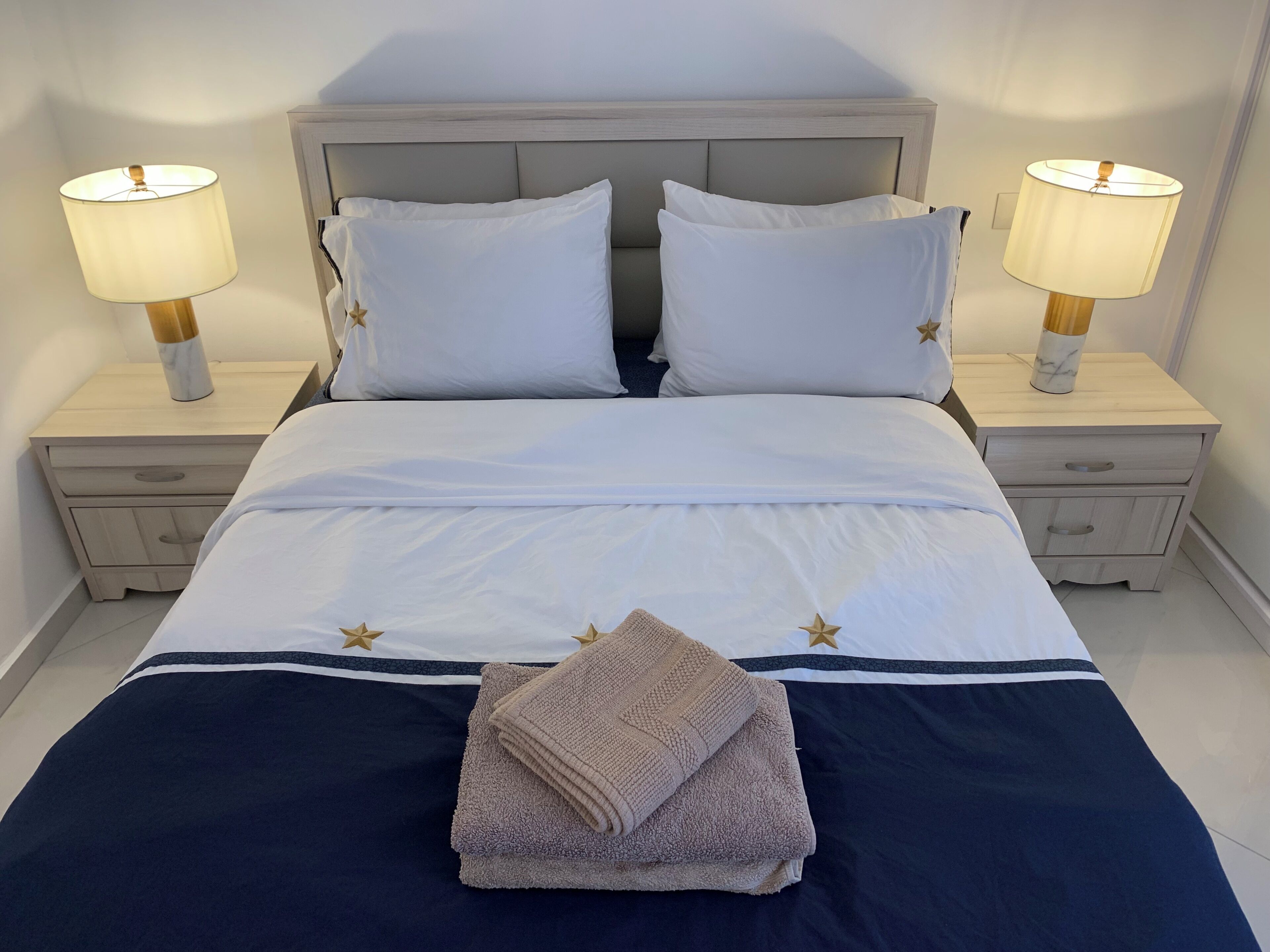 Een keurig opgemaakt bed met witte lakens en twee kussens, aan weerszijden geflankeerd door nachtkastjes met lampen, in een kamer met een strak, minimalistisch design.