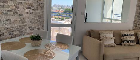 A modern living room with a white table, beige sofa, and a view of buildings through a large window.