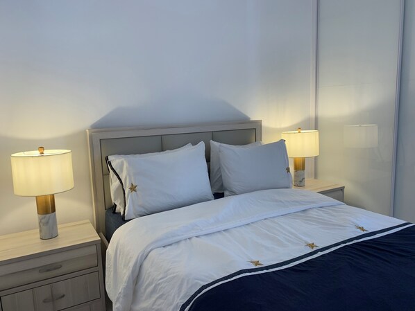 Una cama bien tendida con ropa blanca y almohadas estampadas con estrellas, flanqueada por mesitas de noche con lámparas, en una habitación de diseño sencillo y limpio.