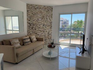 A living room with a beige sofa, a glass coffee table, and a large window with a view of buildings and a balcony.