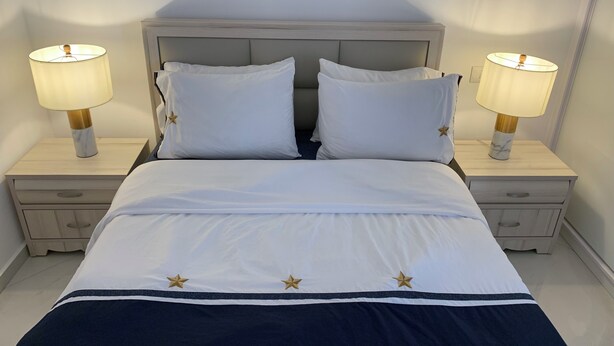 Un lit bien fait, avec du linge de lit blanc et des oreillers à motifs étoilés, flanqué de deux tables de chevet sur lesquelles sont posées des lampes.
