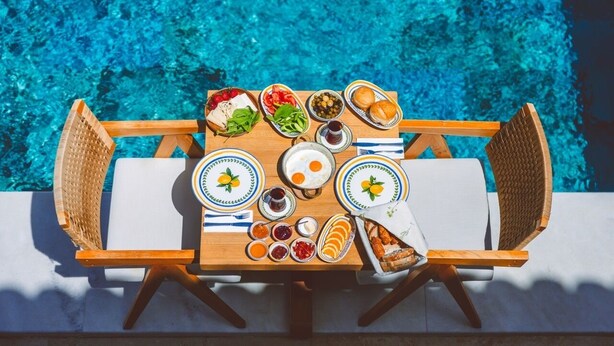 Una mesa de madera con platos de comida, una silla de mimbre y una piscina al fondo.