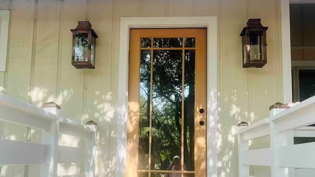 Una terraza de madera que conduce a una puerta blanca con un panel de vidrio, bordeada por barandas blancas y dos faroles.