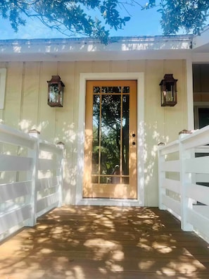 A wooden deck leading to a white door with a glass panel, flanked by white railings and two lanterns.