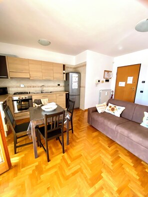 A compact kitchen with a refrigerator, stove, and sink. There is a dining table with chairs and a sofa in the living area. - Luigina's Home • Direct Metro to the Vatican, Terrace & Parking (Roma)