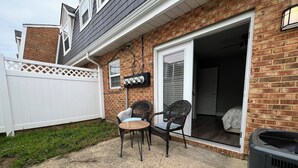 Un patio con dos sillones de mimbre y una mesa, una casa de ladrillo con cerca blanca y un ventanal corredero que da a un dormitorio.