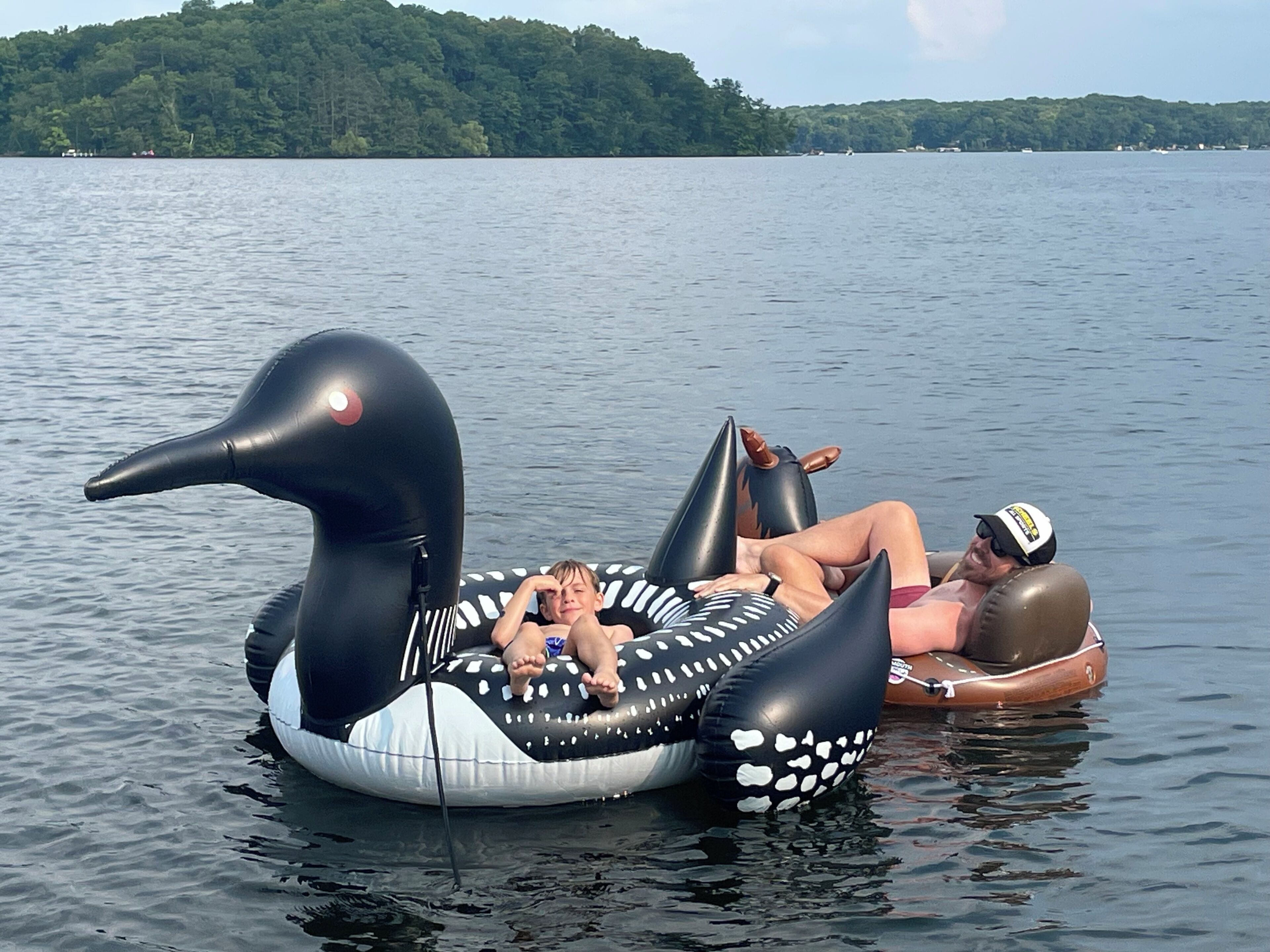 Dos personas sobre un pato inflable gigante flotan en un lago, con una orilla boscosa al fondo.