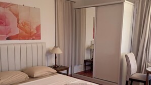Hotelkamer met een bed, een stoel, een bureau, een lamp, een spiegel en een groot raam met gordijnen.