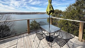 A wooden deck with a metal table and chairs, an umbrella, and a view of a lake and distant buildings. - Wide Sky Chateau Lake Views (Osage Beach)