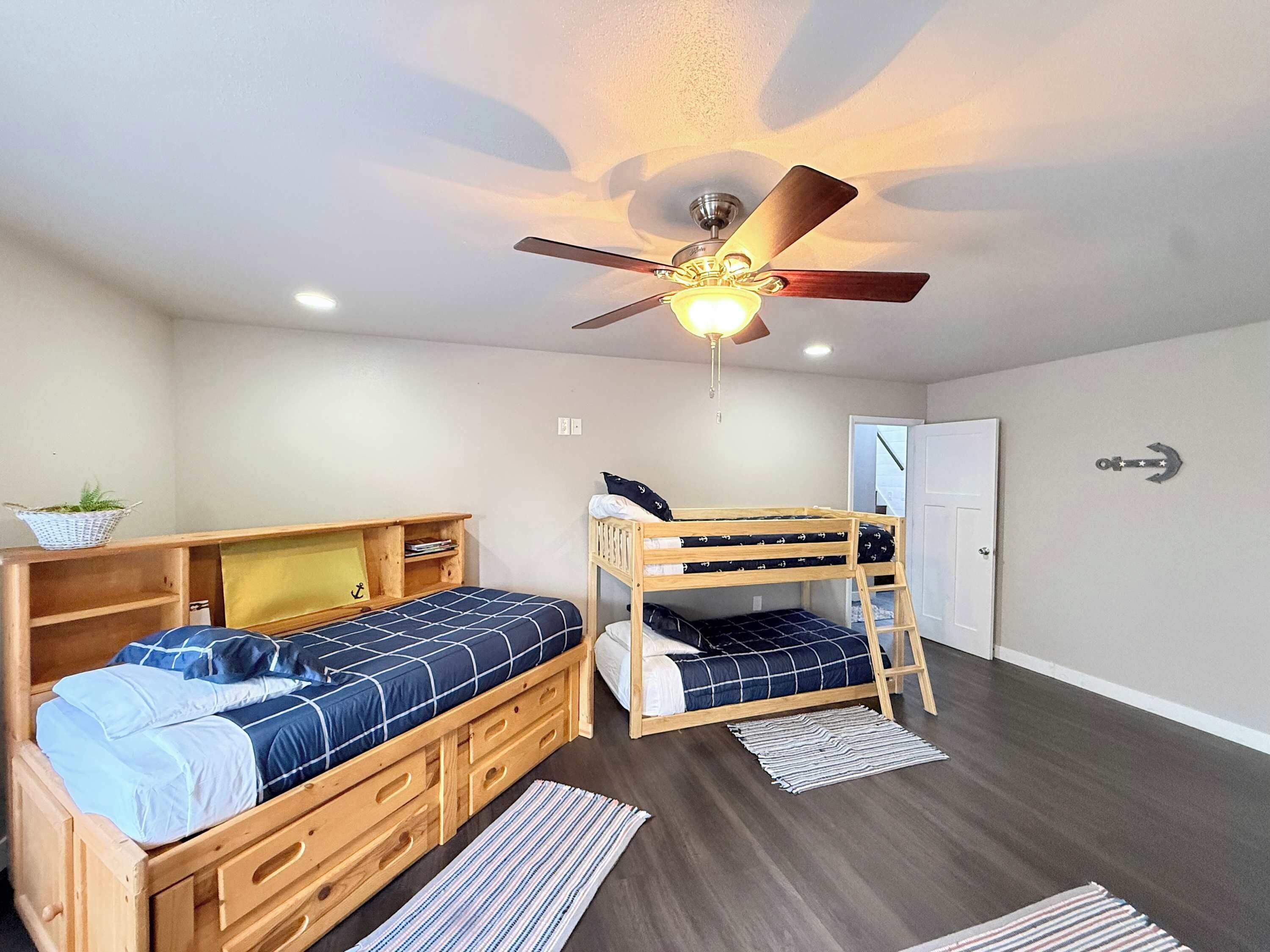 A room with two bunk beds, a ceiling fan, a small shelf with a plant, and a door with an anchor decoration.