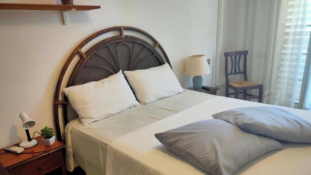 Un lit bien fait, avec une tête de lit, une table de chevet avec une lampe et une petite plante, et une chaise dans une pièce avec une fenêtre.