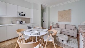 Eine moderne Küche mit weißen Schränken, ein Essbereich mit rundem Tisch und Stühlen sowie ein Wohnbereich mit Sofa und einem Wandbild.