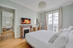 A bedroom with a fireplace, a bed with white bedding, a television above the fireplace, and a view of the outside through a window. - Design Apartment 1br/4p - Monceau (Paris)