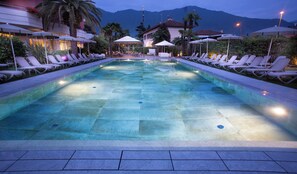 Ein Schwimmbecken mit Liegestühlen und Sonnenschirmen, umgeben von Gebäuden und Bergen.