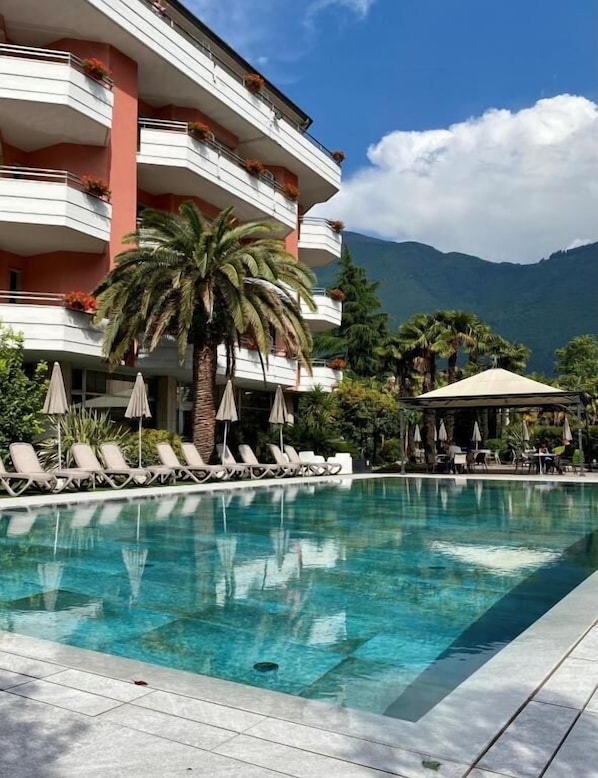 A hotel with a pool, lounge chairs, umbrellas, and a mountain view.