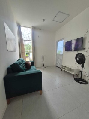 Un salon avec un canapé de couleur turquoise, un ventilateur sur pied, un téléviseur et une fenêtre avec des rideaux.