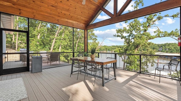 A covered outdoor deck with a wooden table and chairs, overlooking a lake with trees and a distant building.