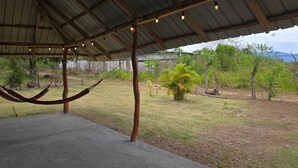 A covered outdoor area with a hammock, concrete floor, and a view of a grassy landscape with trees and mountains in the background.