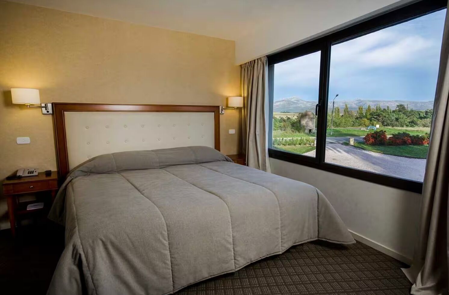 Une chambre d’hôtel avec un lit, une table de chevet, une lampe, une fenêtre donnant sur un paysage verdoyant et des montagnes.