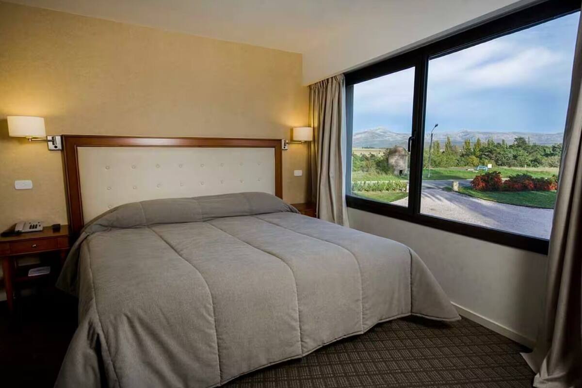 Ein Hotelzimmer mit Bett, Nachttisch, Lampe und Fenster mit Blick auf Grünflächen und Berge.