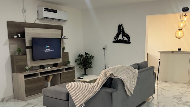 A modern living room with a grey sofa, a flat-screen TV, a wall-mounted air conditioner, and a marble-tiled floor. - Casa completa 2 habitaciones 2 baños a 7 minutos del aeropuerto y 5 de la ciudad (Guaymallen)