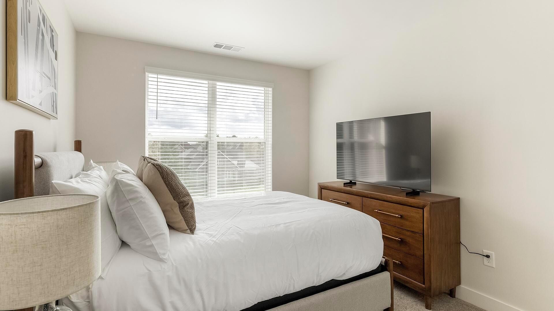 A bedroom with a bed, a TV on a wooden dresser, a window with blinds, and a lamp.