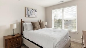 A bedroom with a bed, nightstands, a lamp, a painting, and a window with blinds. - Landing Apartments Huntsville near Lake Forest (Huntsville)