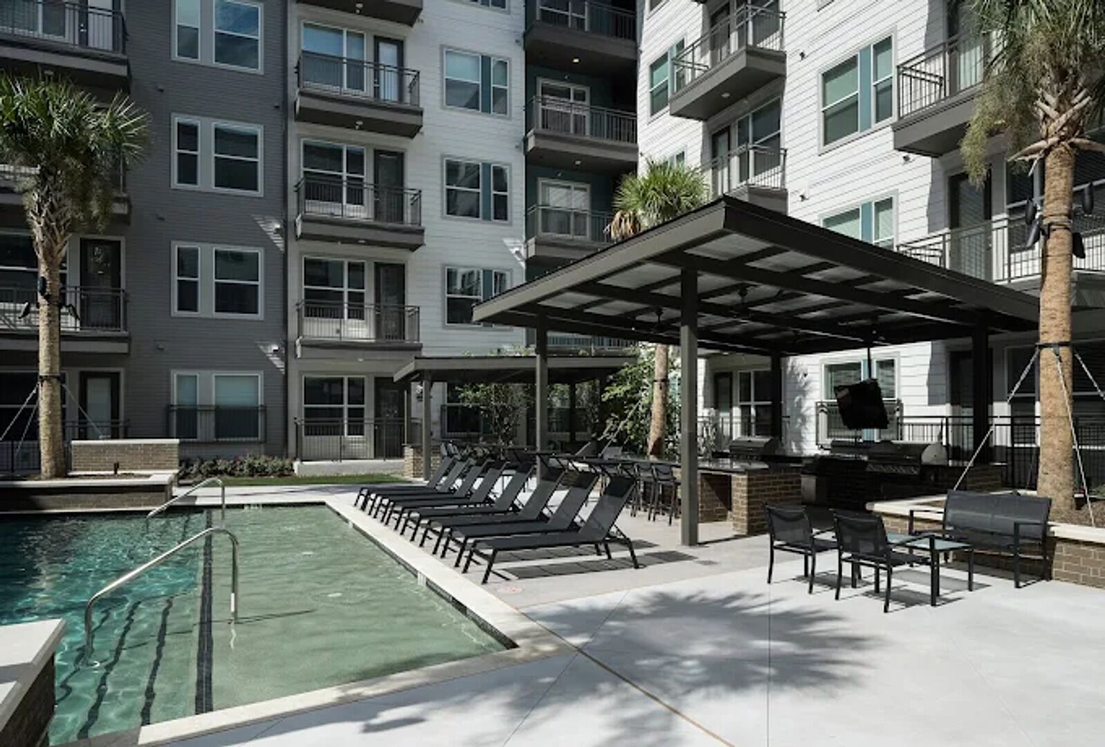 Ein Poolbereich mit Liegestühlen, ein überdachter Grillplatz und ein Gebäude mit mehreren Balkonen.
