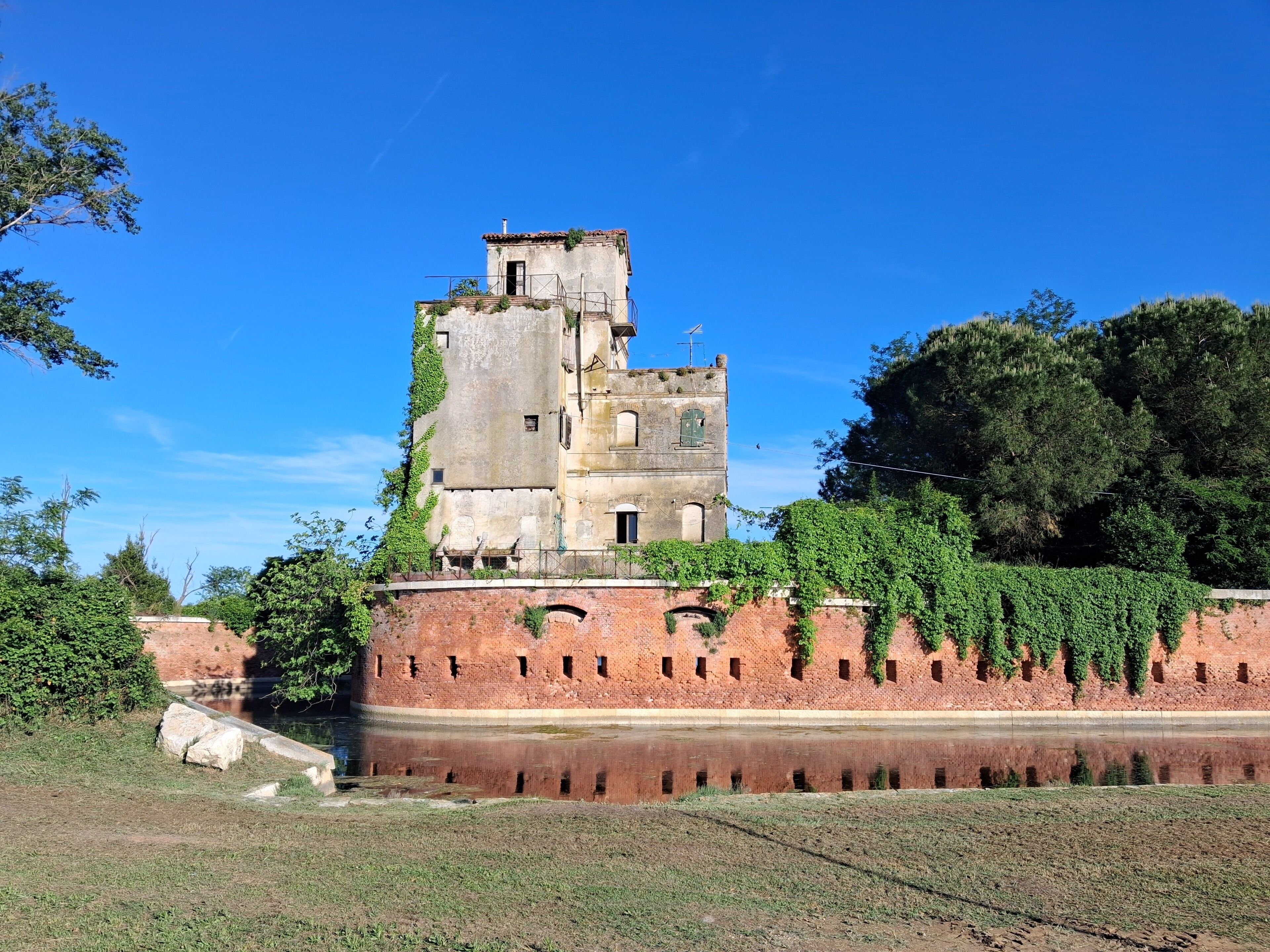 A historic building with a brick wall and a tower, surrounded by greenery and a reflective water body.