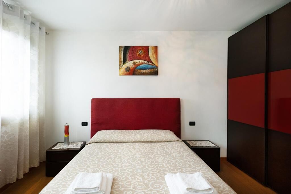A hotel room with a red headboard, a patterned bedspread, a painting on the wall, and a nightstand with a candle.