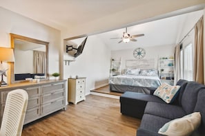 Ein Schlafzimmer mit einem Bett, einem Schreibtisch, einem Stuhl, einer Kommode, einem Deckenventilator und einem Fenster mit Vorhängen.